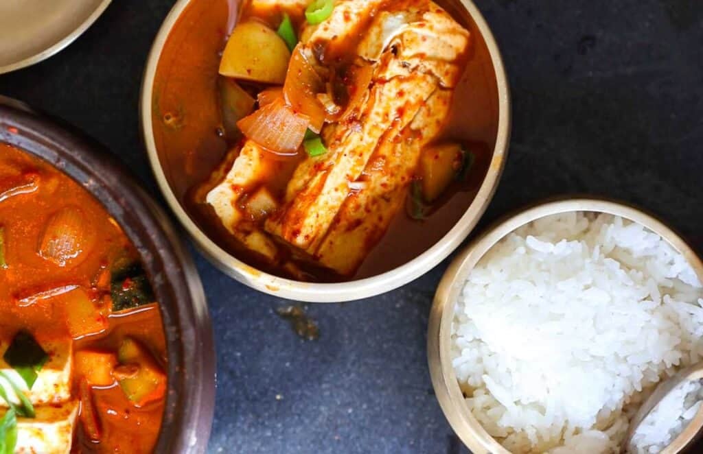 A bowl of spicy doenjang jjigae with tofu, zucchini, onions, and other vegetables is next to a bowl of white rice on a dark surface. Another partial bowl of stew is visible, suggesting a Korean meal setting.